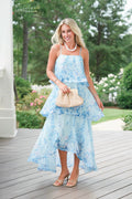 Woman in a light blue floral dress standing on a wooden deck with a garden background