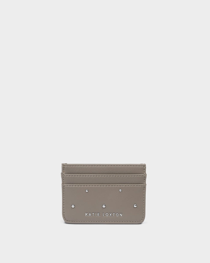 A small taupe leather card holder wallet by Katie Loxton, featuring silver-tone metal studs and the brand name printed in white at the bottom center, set against a solid light gray background.