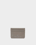 A small taupe leather card holder wallet by Katie Loxton, featuring silver-tone metal studs and the brand name printed in white at the bottom center, set against a solid light gray background.