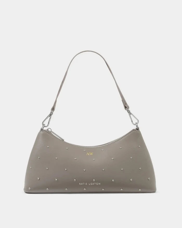 A taupe-brown studded shoulder bag by Katie Linton, featuring a crescent-shaped silhouette, a single shoulder strap, and silver-tone studs across the surface, set against a plain light gray background.