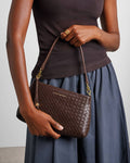 A model holds a brown woven leather shoulder bag with gold hardware and a tassel charm. The model is wearing a dark brown top and a slate blue skirt against a light gray background.