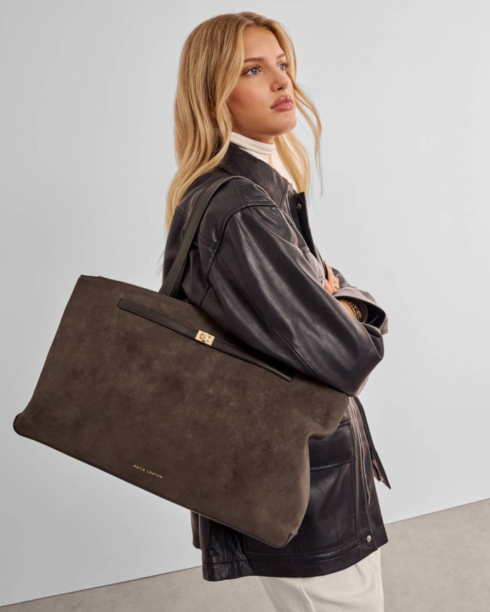 A woman wearing a black leather jacket and cream turtleneck carries a large dark brown suede tote bag with a black leather strap and gold hardware.