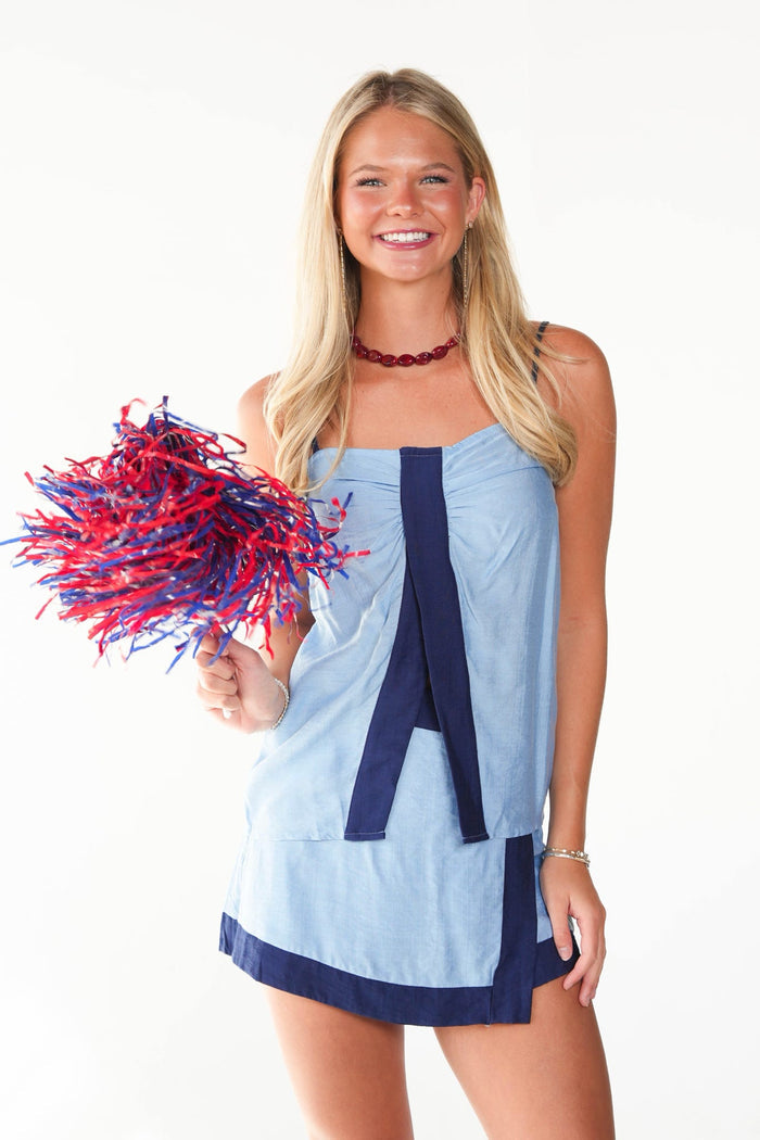 A smiling blonde woman stands against a white studio background wearing a light blue and navy mini dress and holding a red and blue pom-pom.