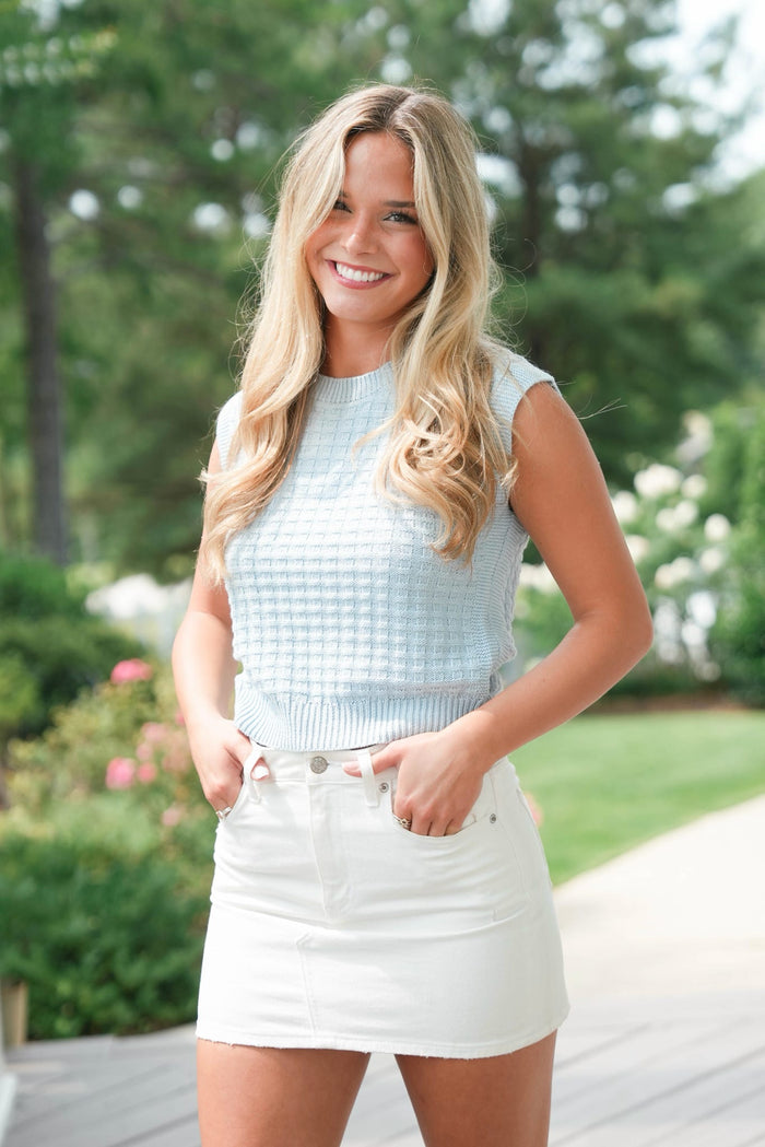 Woman in light blue sleeveless top and white skirt standing outdoors with greenery in the background