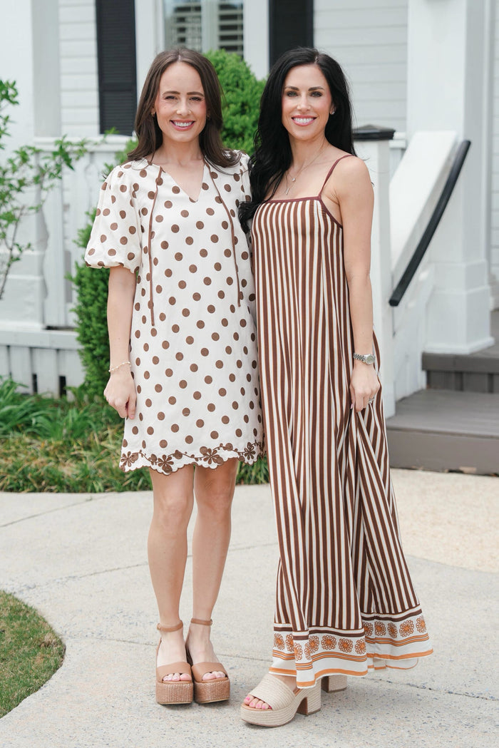 Two women standing outdoors, one in a polka dot dress and the other in a striped jumpsuit.