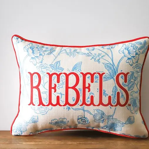 White pillow with blue florals and red "Rebels" with red piping around the edge. On a white background.