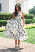 A woman wearing a sleeveless white midi dress with a black and gray botanical print, standing outdoors on a paved walkway in front of a white building and green hedges.