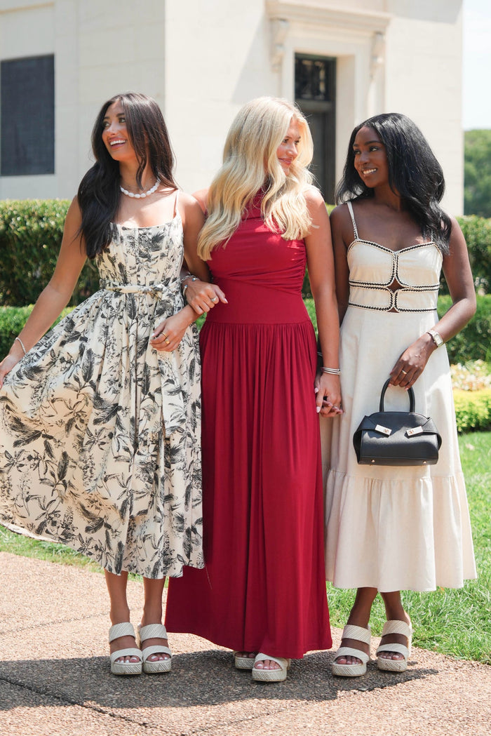Three women standing outdoors in front of a white building, each wearing a different formal dress. The woman on the left wears a sleeveless beige and black floral midi dress, the woman in the center wears a solid deep red sleeveless maxi dress, and the woman on the right wears a cream strapless midi dress with gold trim and carries a black handbag.