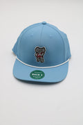 A light blue flat-brim cap with a white piping detail and a central embroidered patch of a cartoon bear holding a red heart. A green circular sticker is visible on the brim.