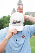 A person outdoors wearing a white baseball cap with 'Tigers Auburn' embroidered on the front, paired with a light blue patterned polo shirt. A red and white Auburn logo is visible on the chest, and a brick clock tower is blurred in the background.