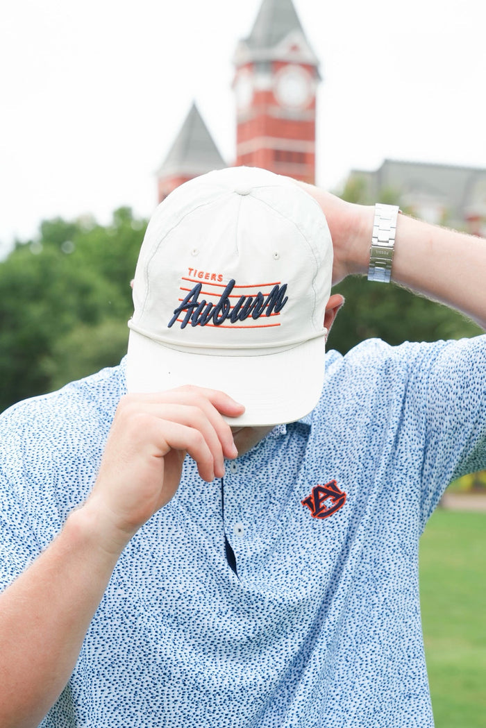 A person outdoors wearing a white baseball cap with 'Tigers Auburn' embroidered on the front, paired with a light blue patterned polo shirt. A red and white Auburn logo is visible on the chest, and a brick clock tower is blurred in the background.