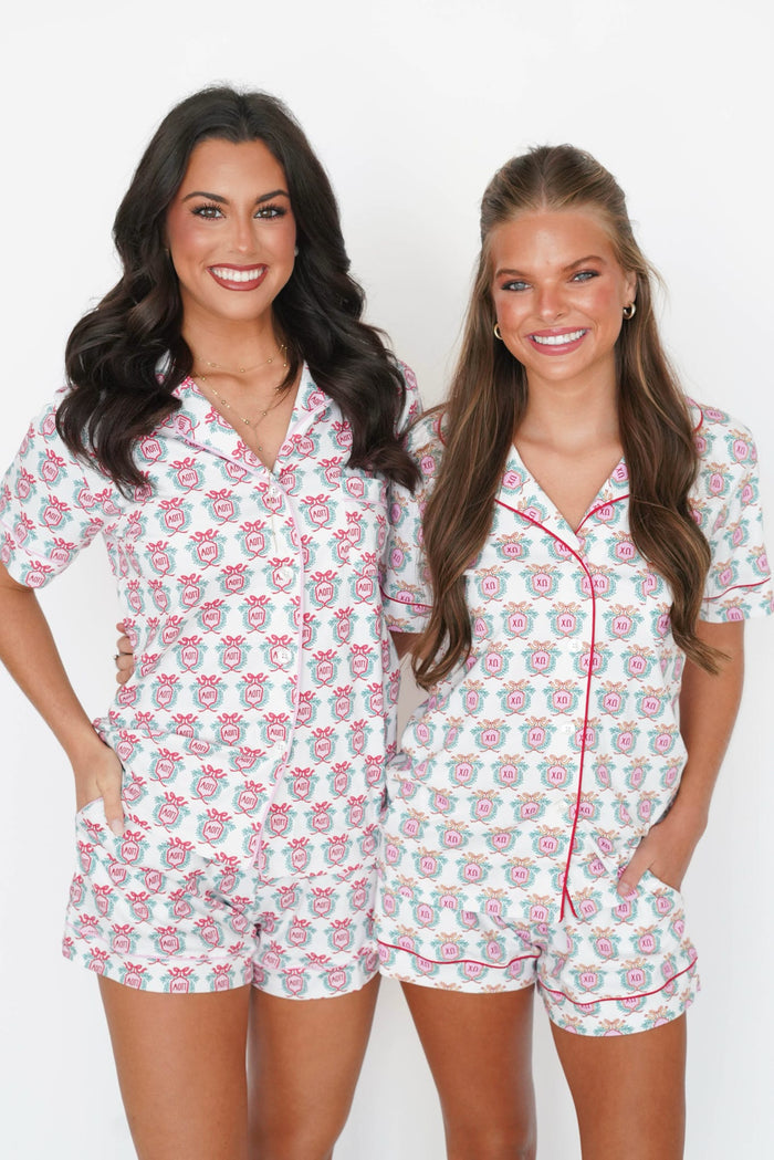 Two women modeling matching white short-sleeve pajama sets with a pink and gray floral pattern and red piping, standing against a plain white background.