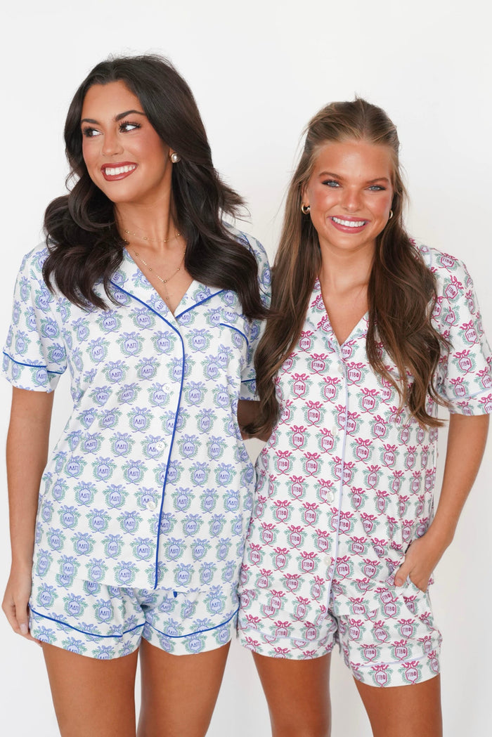 Two women modeling matching short-sleeve pajama sets against a plain white background. The set on the left is white with a blue floral pattern and blue piping, and the set on the right is white with a pink floral pattern and pink piping.