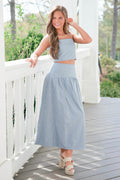 Woman in a light blue striped outfit standing on a wooden deck with greenery in the background