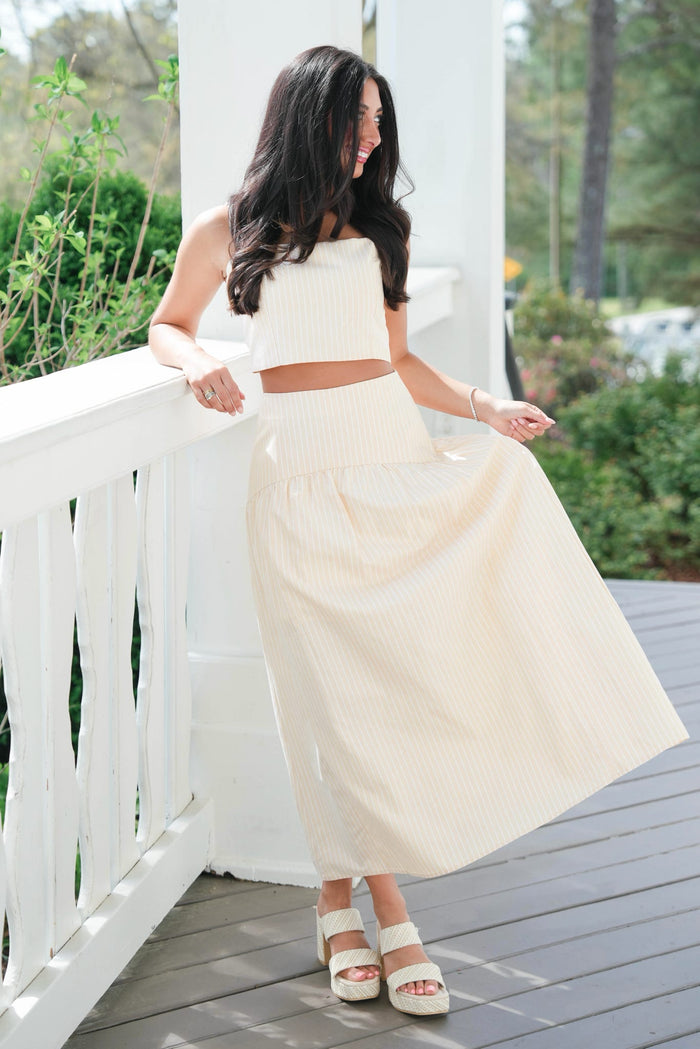 Woman in a white top and cream skirt standing on a wooden deck with greenery in the background