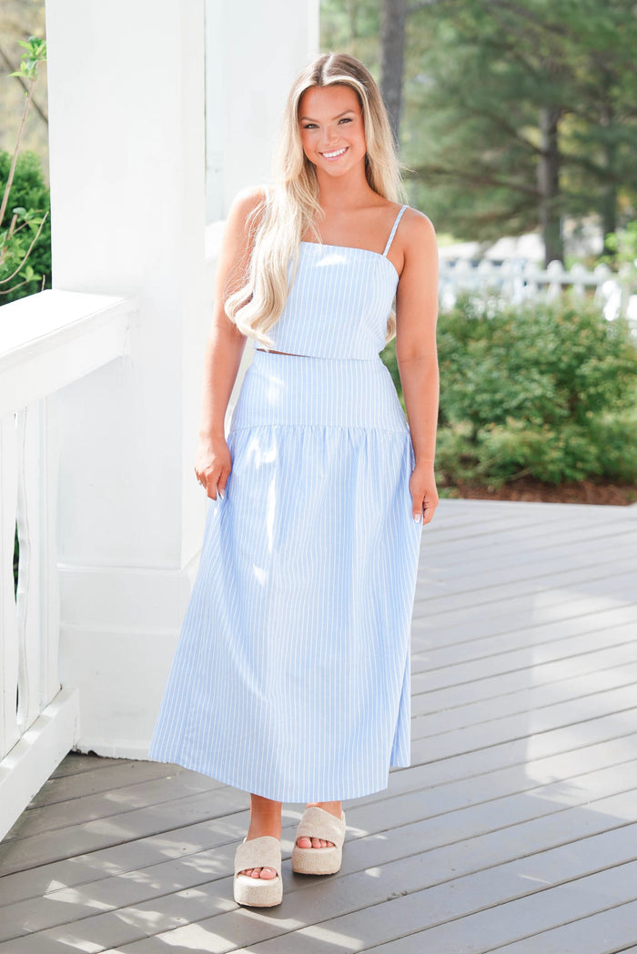 Woman wearing a light blue dress standing on a wooden deck with greenery in the background