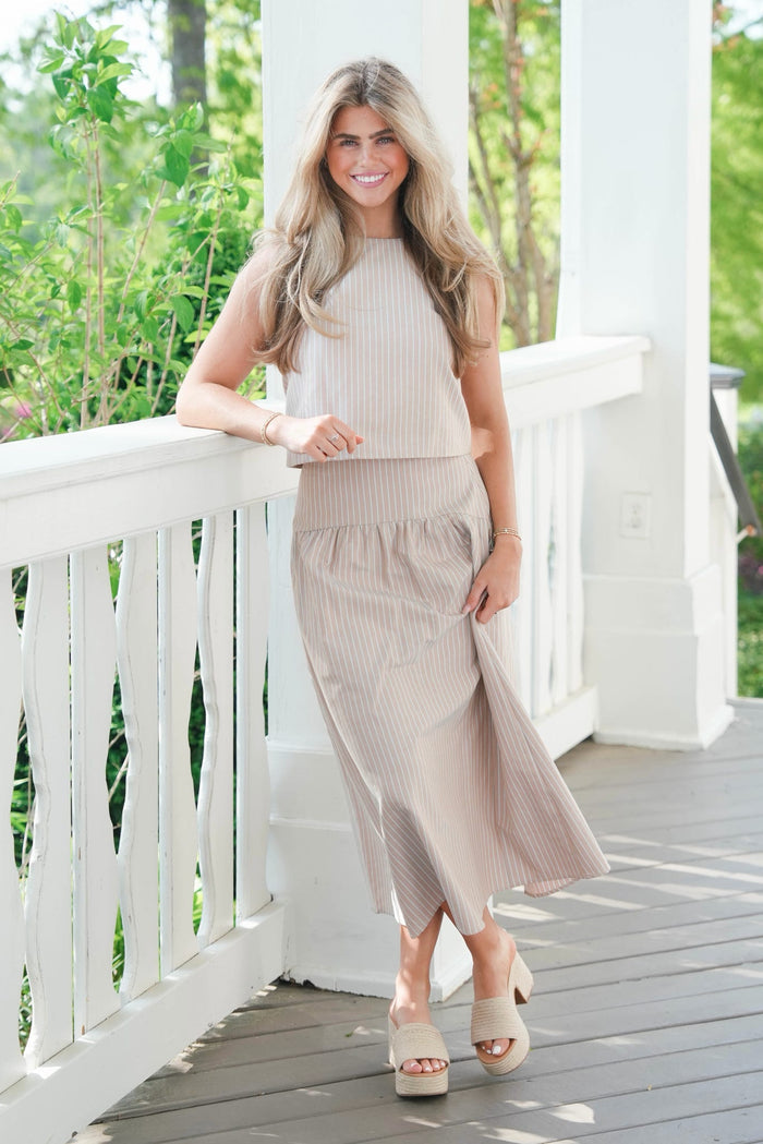 Woman in a beige sleeveless top and skirt standing on a wooden deck with greenery in the background
