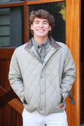 A smiling young man wearing a light beige diamond-quilted jacket over a green plaid shirt and white pants, standing in front of a rustic wooden door.