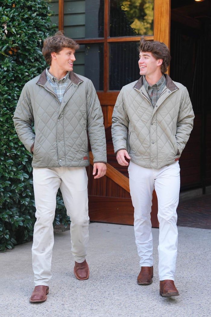Two men walking outdoors wearing matching quilted light olive green jackets with brown collar trim, paired with light pants and brown shoes, in front of a rustic wooden building.