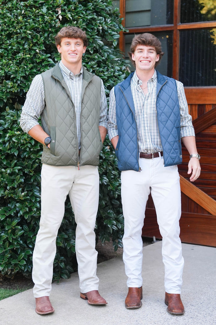 Two men standing outdoors in front of a green hedge and wooden door, each wearing a quilted vest over a plaid button-up shirt and white pants.