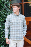 A man wearing a light gray and white plaid long-sleeve button-down shirt with beige pants, standing outdoors in front of a wooden door and green foliage.