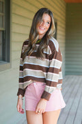 A woman wearing a brown, beige, and white striped knit sweater and a pink mini skirt, standing on a wooden porch.