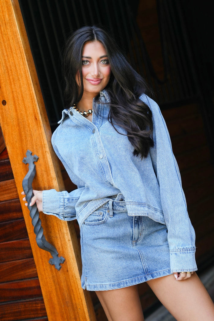 A woman wearing a light blue denim long-sleeve button-up shirt and matching denim mini skirt, leaning against a wooden door frame.