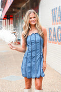 A woman wearing a strapless blue denim-like dress with vertical gathered seams, standing outdoors on a sidewalk next to a white brick wall with large lettering. She is holding a white feather boa.