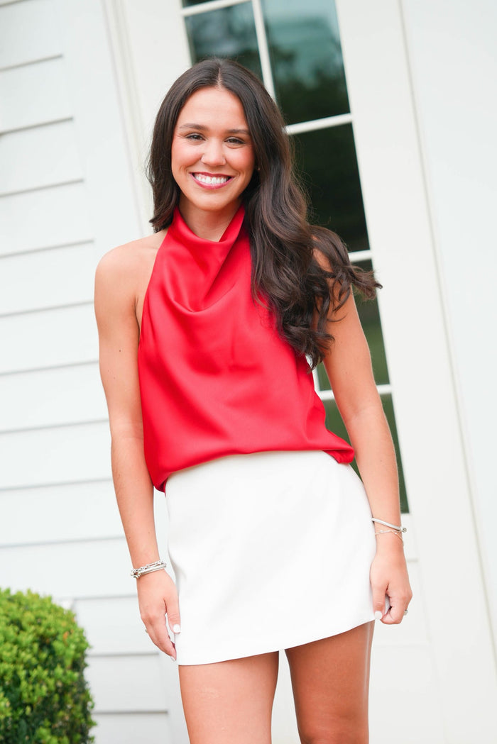 Woman wearing a red top and white skirt standing in front of a white door.