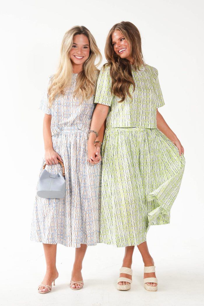 Two women modeling matching short-sleeve midi dresses with a vertical abstract stripe pattern. The woman on the left wears a light blue, white, and pink patterned dress and holds a small gray handbag.