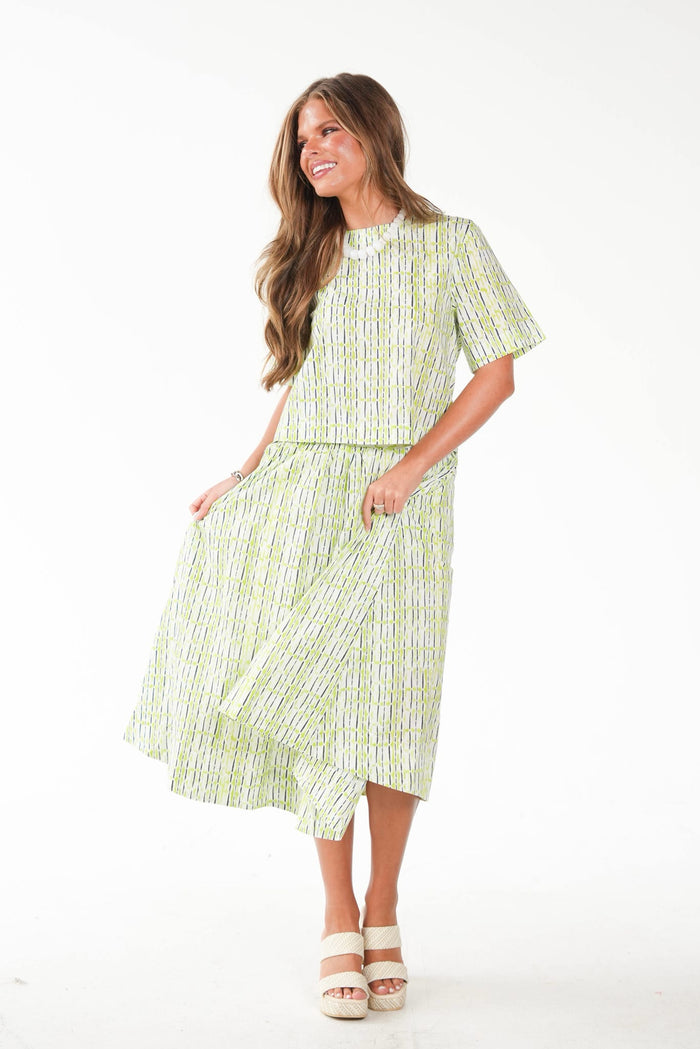 A woman modeling a light green and white vertically striped two-piece set consisting of a short-sleeve top and a matching midi skirt with pockets, paired with white platform sandals, against a plain white studio background.