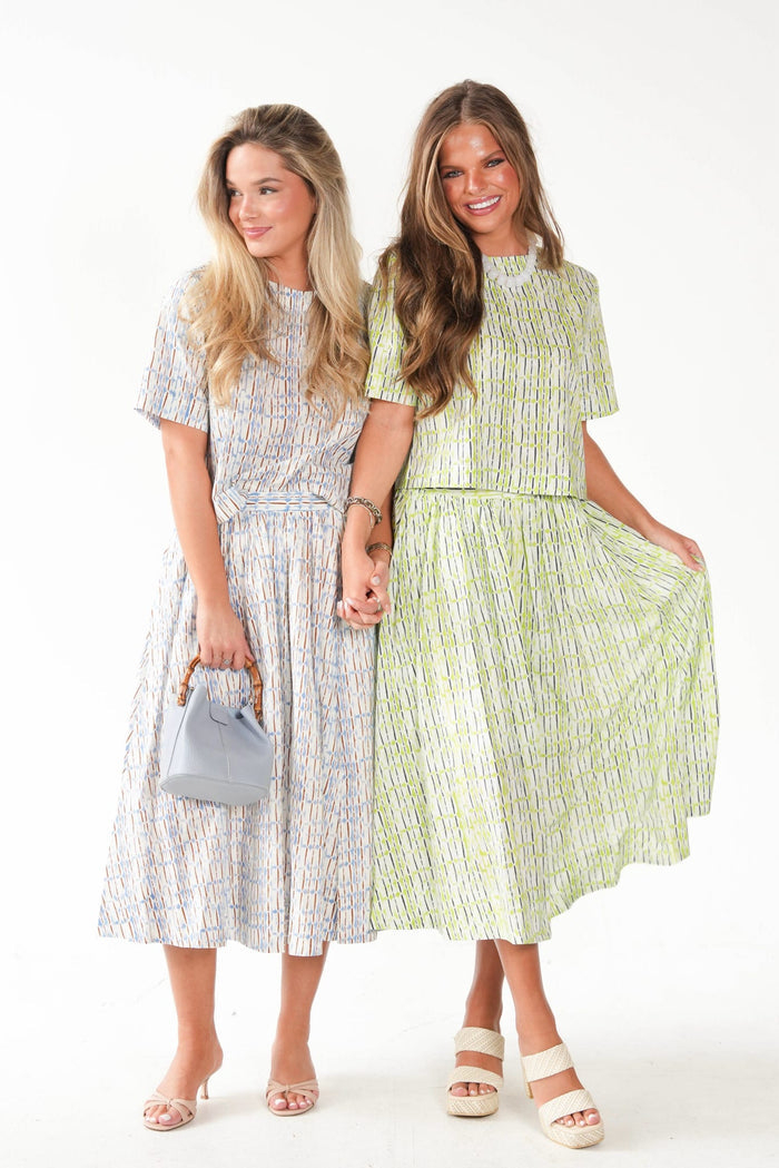 Two women modeling matching midi dresses with a vertical abstract pattern. The dress on the left is light blue, white, and gray, while the dress on the right is light green and white.