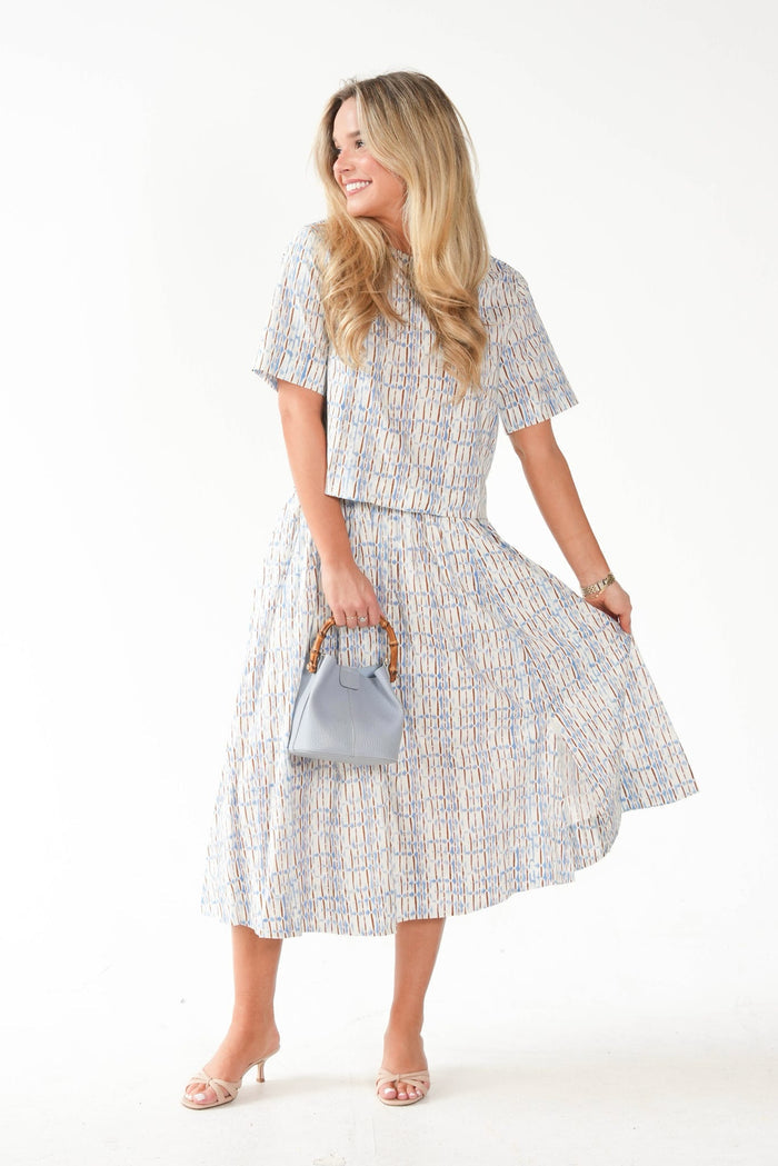 A woman modeling a matching short-sleeve top and midi skirt set in a light blue, white, and beige geometric pattern, holding a small light blue handbag against a plain white studio background.
