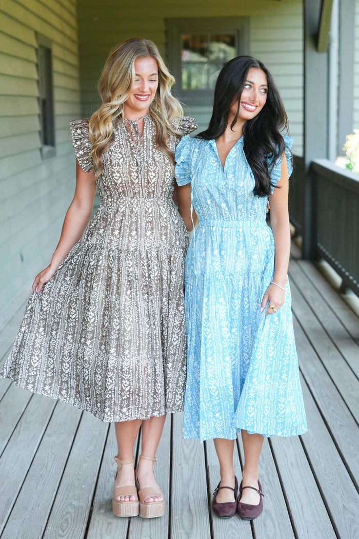 Two women standing on a wooden porch wearing midi-length patterned dresses, one in a light beige/taupe dress and the other in a light blue dress.
