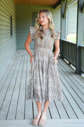 A woman stands on a wooden porch wearing a sleeveless, midi-length lace dress with ruffled cap sleeves and a silver and beige floral pattern. She is wearing beige platform sandals.