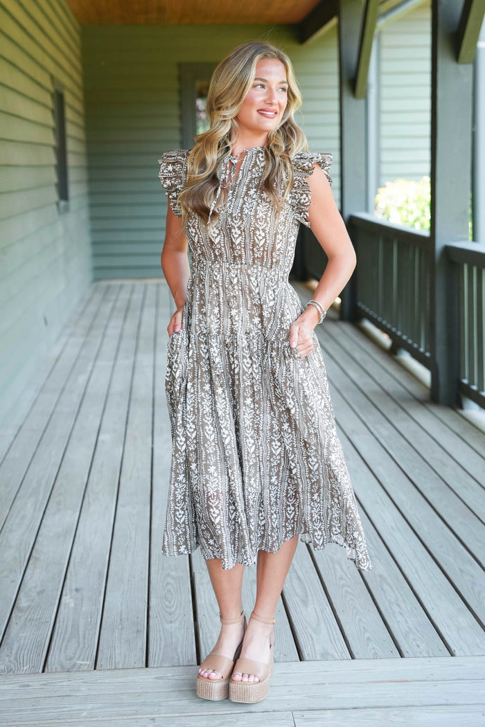 A woman stands on a wooden porch wearing a sleeveless, midi-length lace dress with ruffled cap sleeves and a silver and beige floral pattern. She is wearing beige platform sandals.