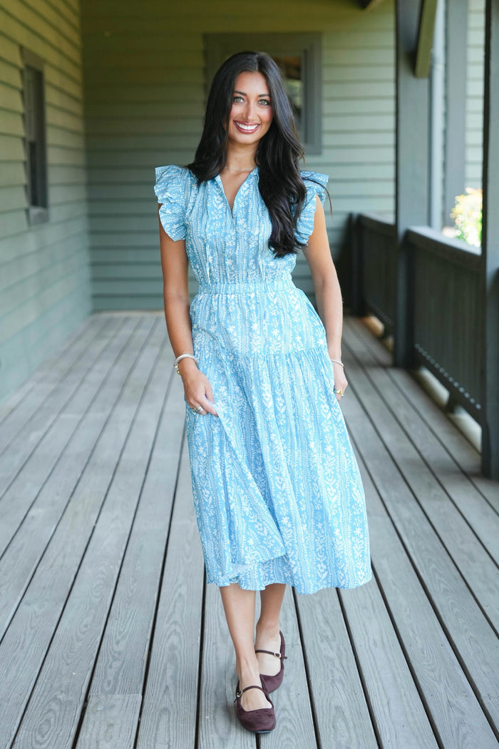 A woman stands on a wooden porch wearing a light blue midi dress with a white floral pattern, ruffled cap sleeves, and a V-neckline.