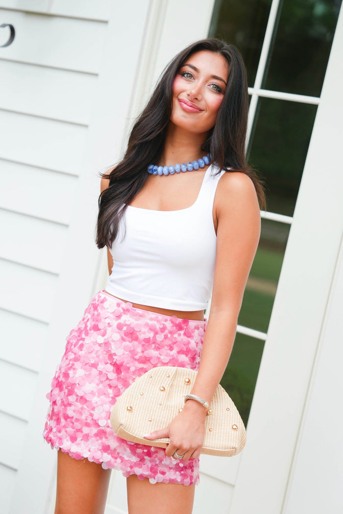Woman wearing a white top and pink floral skirt with a blue necklace, standing against a white wall.