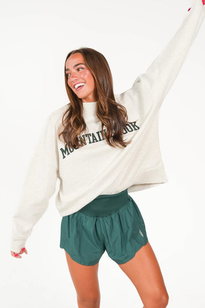 A woman modeling a cream-colored sweatshirt with 'MOUNTAIN VIEW' printed in dark green across the chest, paired with dark green athletic shorts, against a solid white background.