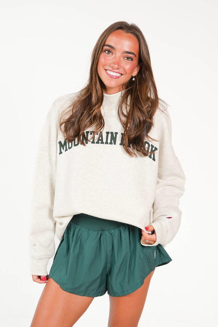 A woman modeling a cream-colored crewneck sweatshirt with 'MOUNTAIN VIEW' printed in dark green across the chest, paired with dark green athletic shorts, against a plain white background.