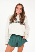 A woman modeling a cream-colored crewneck sweatshirt with 'MOUNTAIN VIEW' printed in dark green across the chest, paired with dark green athletic shorts, against a plain white background.