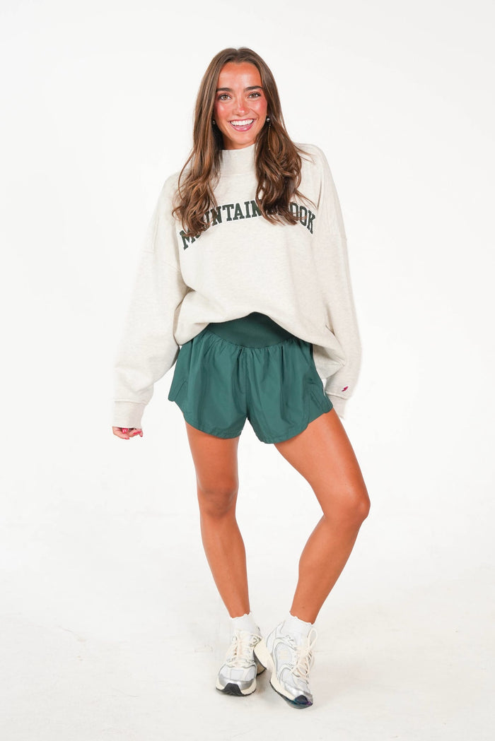 A woman modeling a cream sweatshirt with 'MOUNTAIN VIEW' printed in black across the chest, paired with dark green athletic shorts and white sneakers, against a plain white studio background.