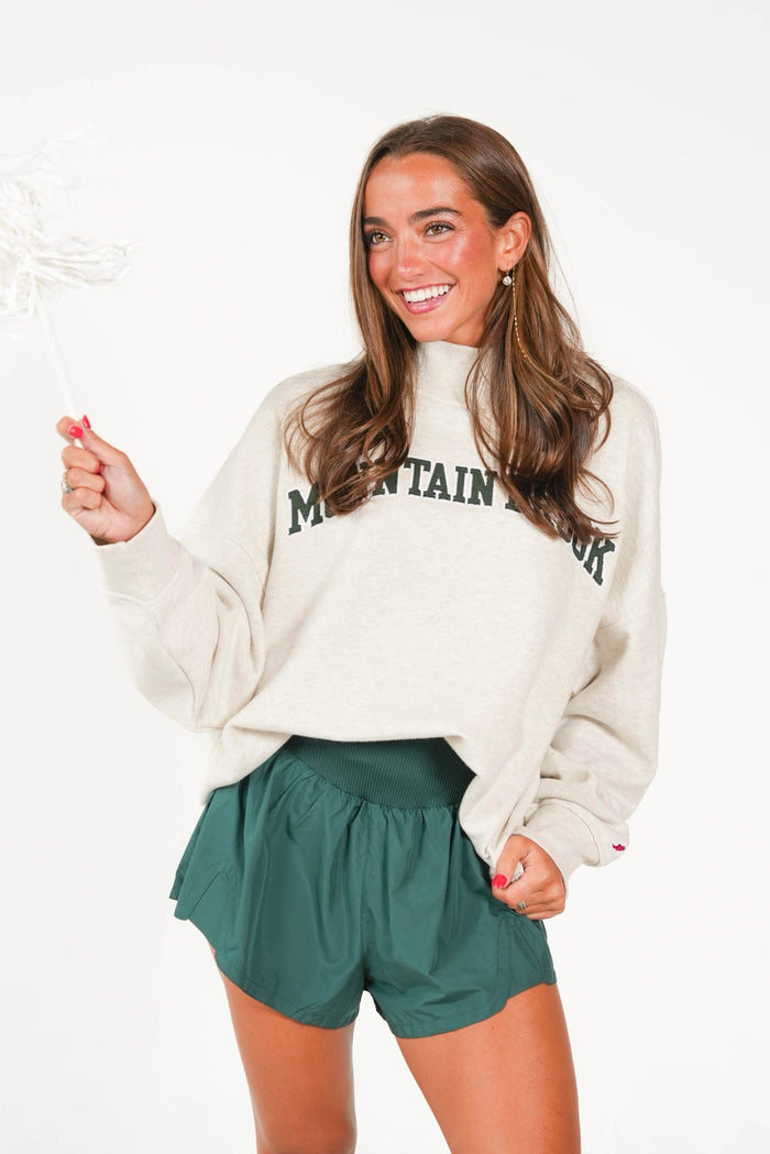 A woman modeling a cream-colored sweatshirt with 'MOUNTAIN VIEW' printed in dark green across the chest, paired with dark green shorts, against a plain white background.