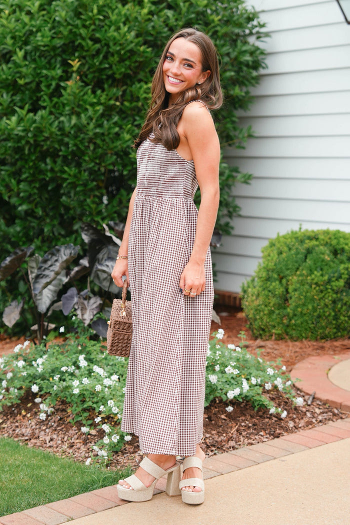 Woman in a checkered dress standing outdoors next to a house with greenery.