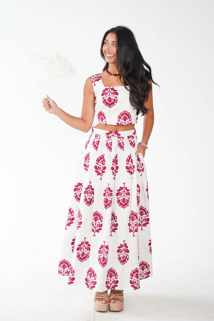 A woman modeling a two-piece white and magenta block-print outfit consisting of a sleeveless crop top and a matching maxi skirt, holding a white paper fan against a plain white background.