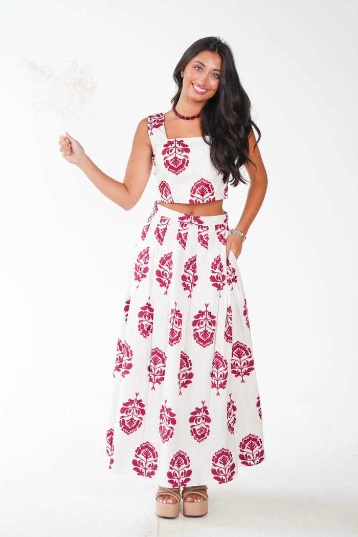 A woman modeling a sleeveless white two-piece set featuring a red and pink block print pattern, consisting of a crop top and a matching maxi skirt, against a plain white studio background.