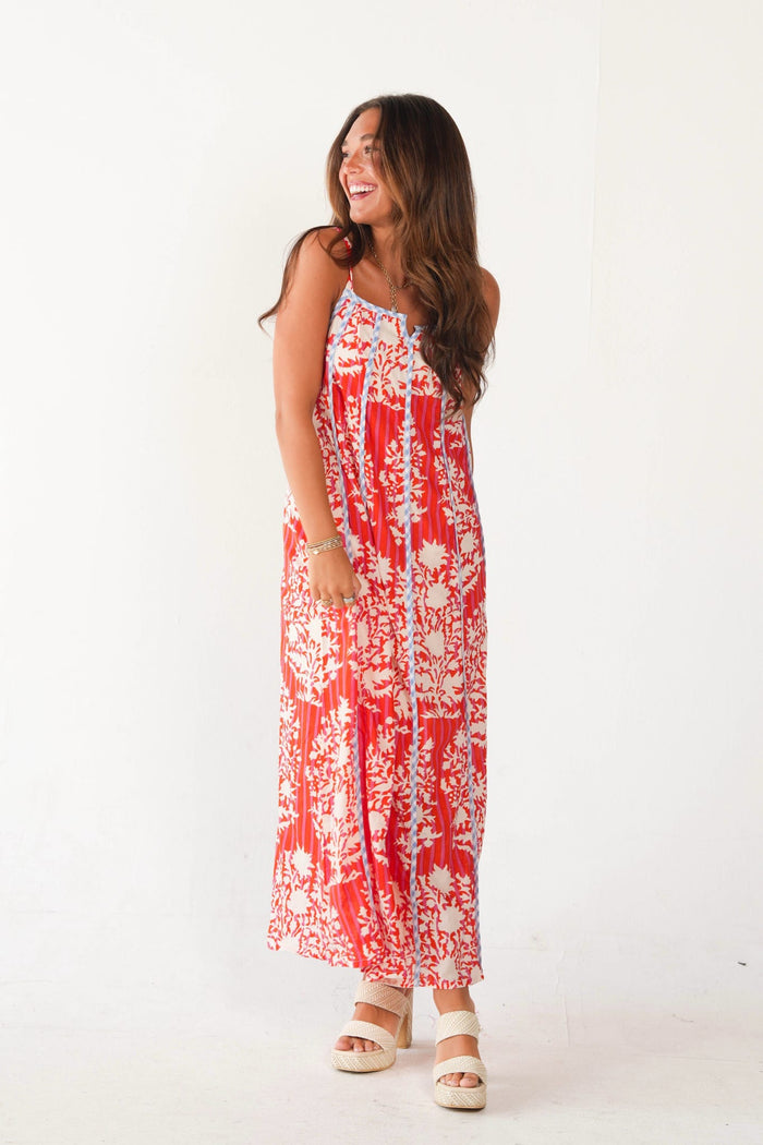 A woman modeling a sleeveless red and white floral maxi dress with a tie-front closure, paired with beige platform sandals, against a plain white studio background.