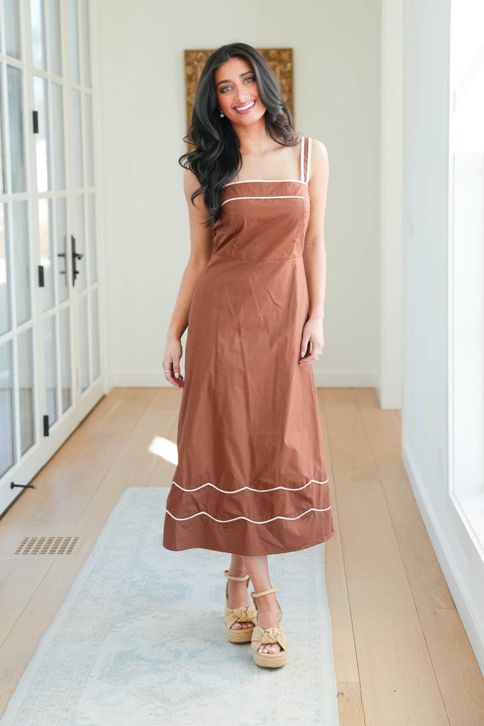 Woman wearing a brown dress with white trim in a room with wooden floor and white walls.