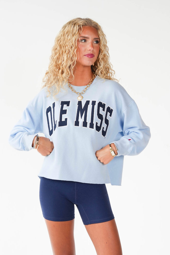 A woman modeling a light blue crewneck sweatshirt with 'OLE MISS' printed in navy blue across the chest, paired with navy blue bike shorts, against a plain white studio background.