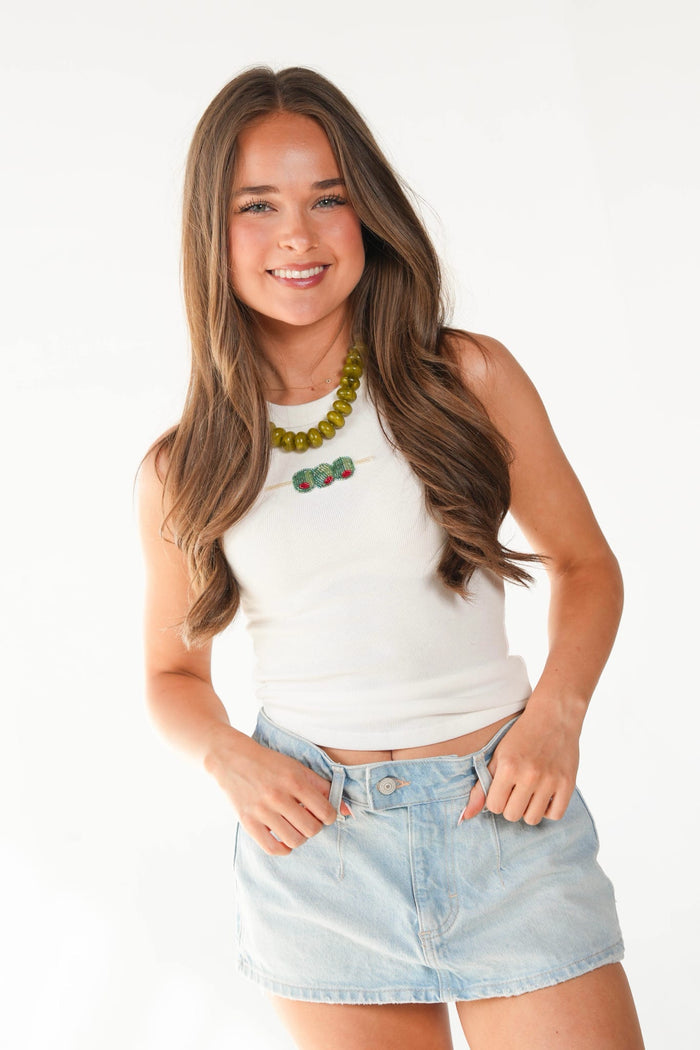 A smiling young woman with long brown hair models a white ribbed tank top and a light blue denim mini skirt against a plain white studio background. She is also wearing a chunky green beaded necklace.